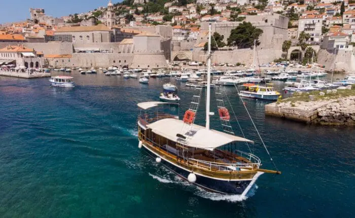 A wooden boat sails in clear blue waters near the historic coastal town of Dubrovnik, with many other boats docked at the marina and houses nestled on a hill in the background. The stunning scene invites you to explore Private Boat Tours or embark on unforgettable Day Trips.