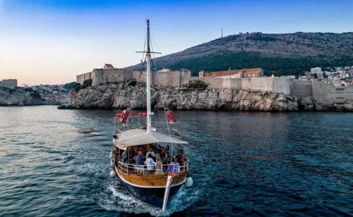 A wooden boat with passengers sails on the calm water near the historic coastal city of Dubrovnik, with stone buildings and a hill in the background, offering idyllic private boat tours and day trips.