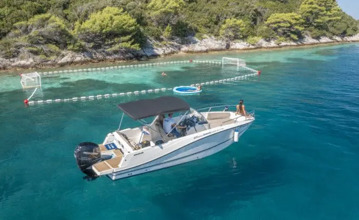 A private boat tour is anchored in clear blue water near a rocky coastline with trees. A person is standing on the boat, and floating water polo goals are visible in the background, making for an unforgettable day trip from Dubrovnik.