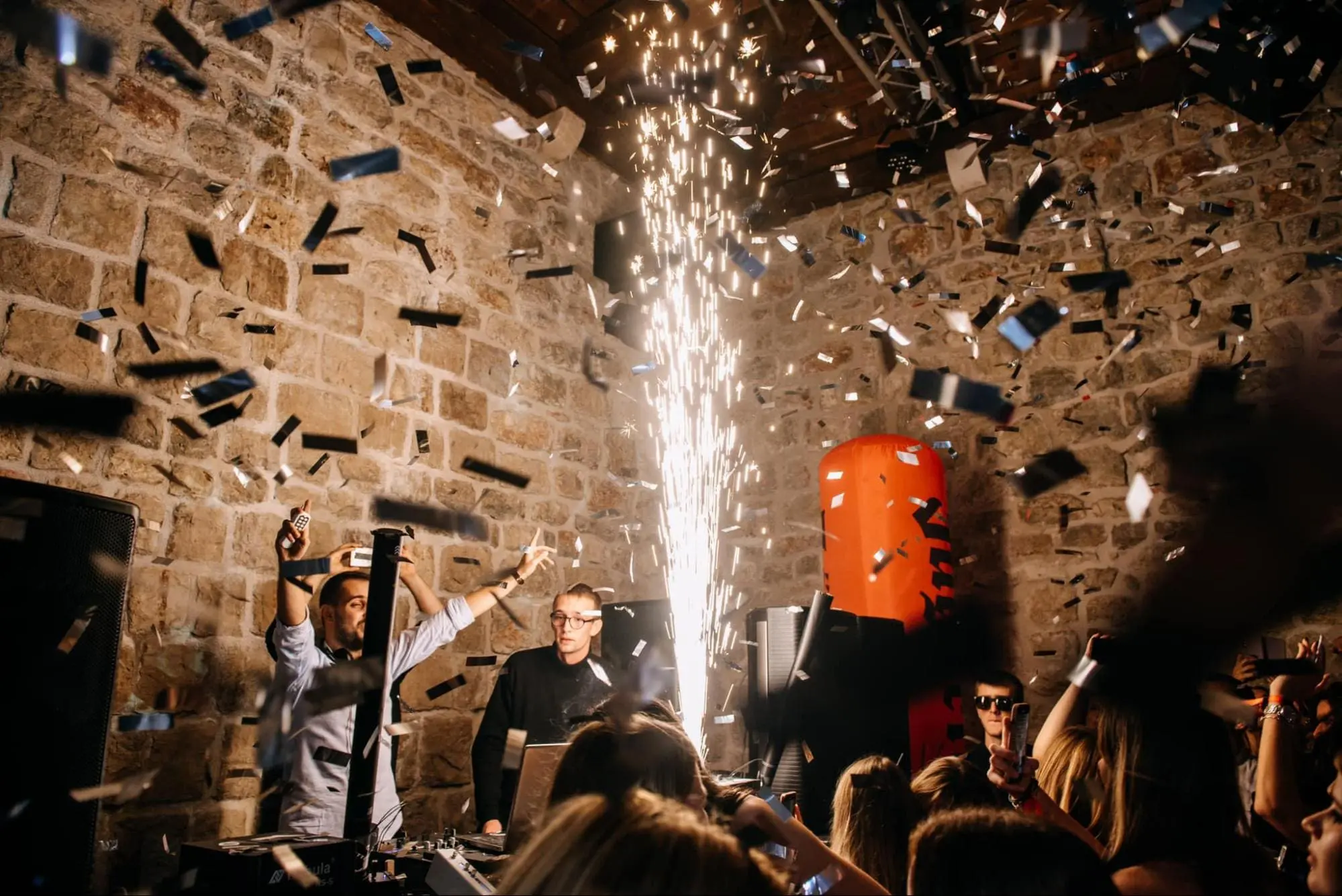 A DJ set is performed at a lively indoor party in Dubrovnik, with two DJs on stage, confetti falling, and a burst of sparks creating an energetic atmosphere. The vibe rivals that of the best day trips or private boat tours along the stunning Adriatic coast.