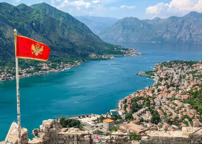 A red flag with a golden emblem on a pole overlooks Dubrovnik, a coastal town and bay surrounded by mountains. The buildings cluster near the water while lush green hills rise in the background, making it an ideal spot for day trips or private boat tours.