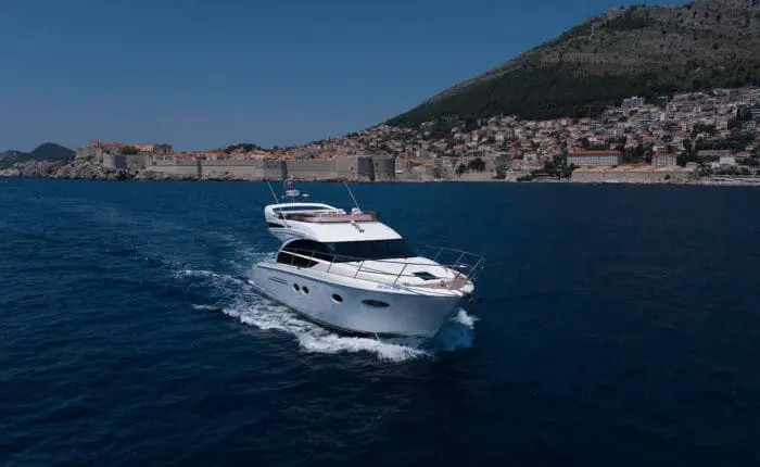 A white yacht cruises on clear blue waters near Dubrovnik, with hillside houses and a historic fortification wall in the background. Perfect for private boat tours or day trips, this scenic coastal town offers an idyllic escape.