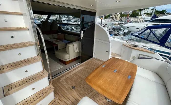 The deck of a modern yacht with wooden flooring and a small table leads to an interior seating area through sliding glass doors, perfect for private boat tours. Ideal for day trips around Dubrovnik, a staircase on the left ascends to an upper level.