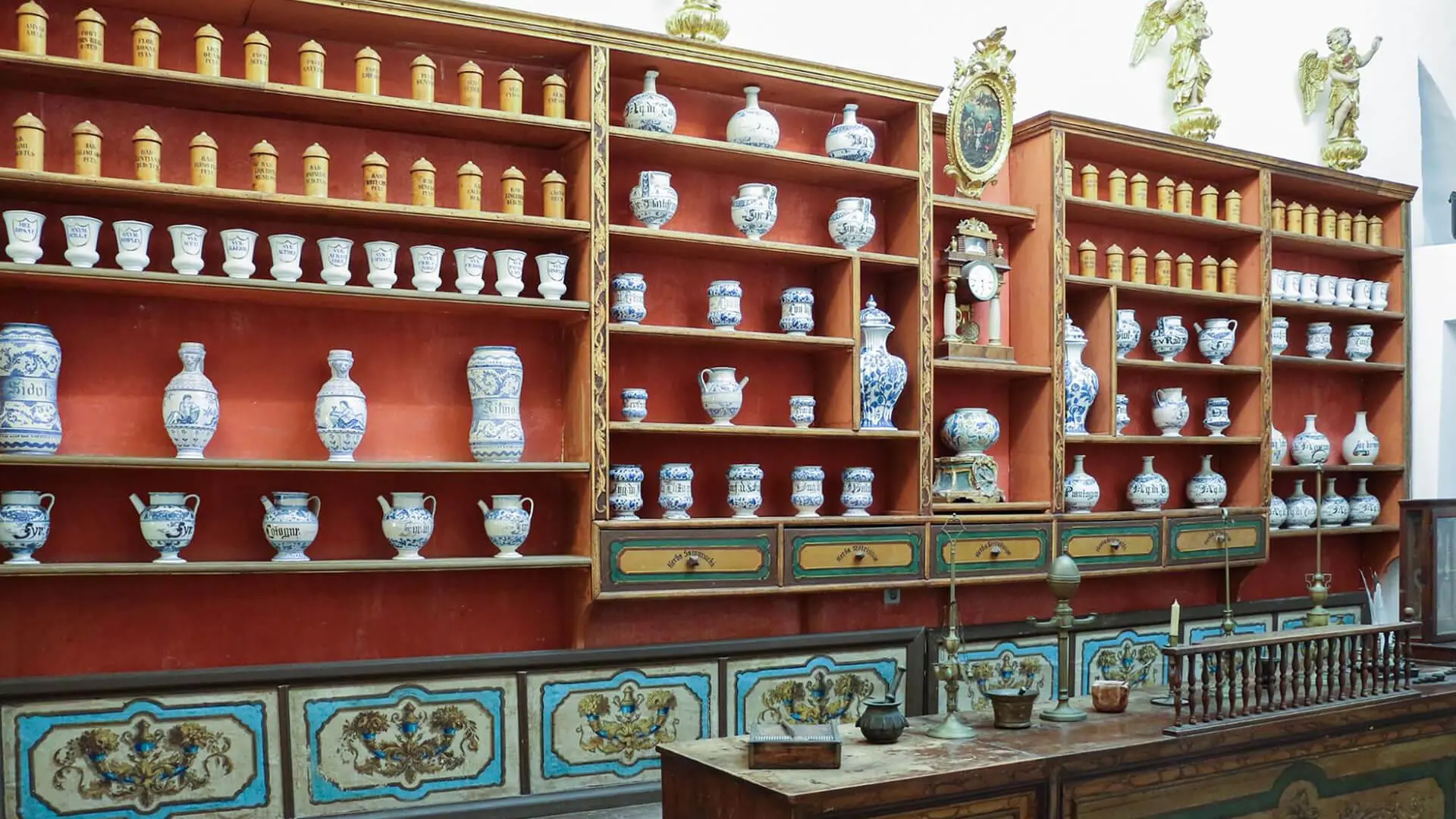 A historical apothecary shelf with blue and white ceramic jars and containers, some labeled, alongside wooden drawers and decorative accents, viewed from the front in a well-lit room—perfect for those interested in Dubrovnik day trips or private boat tours.