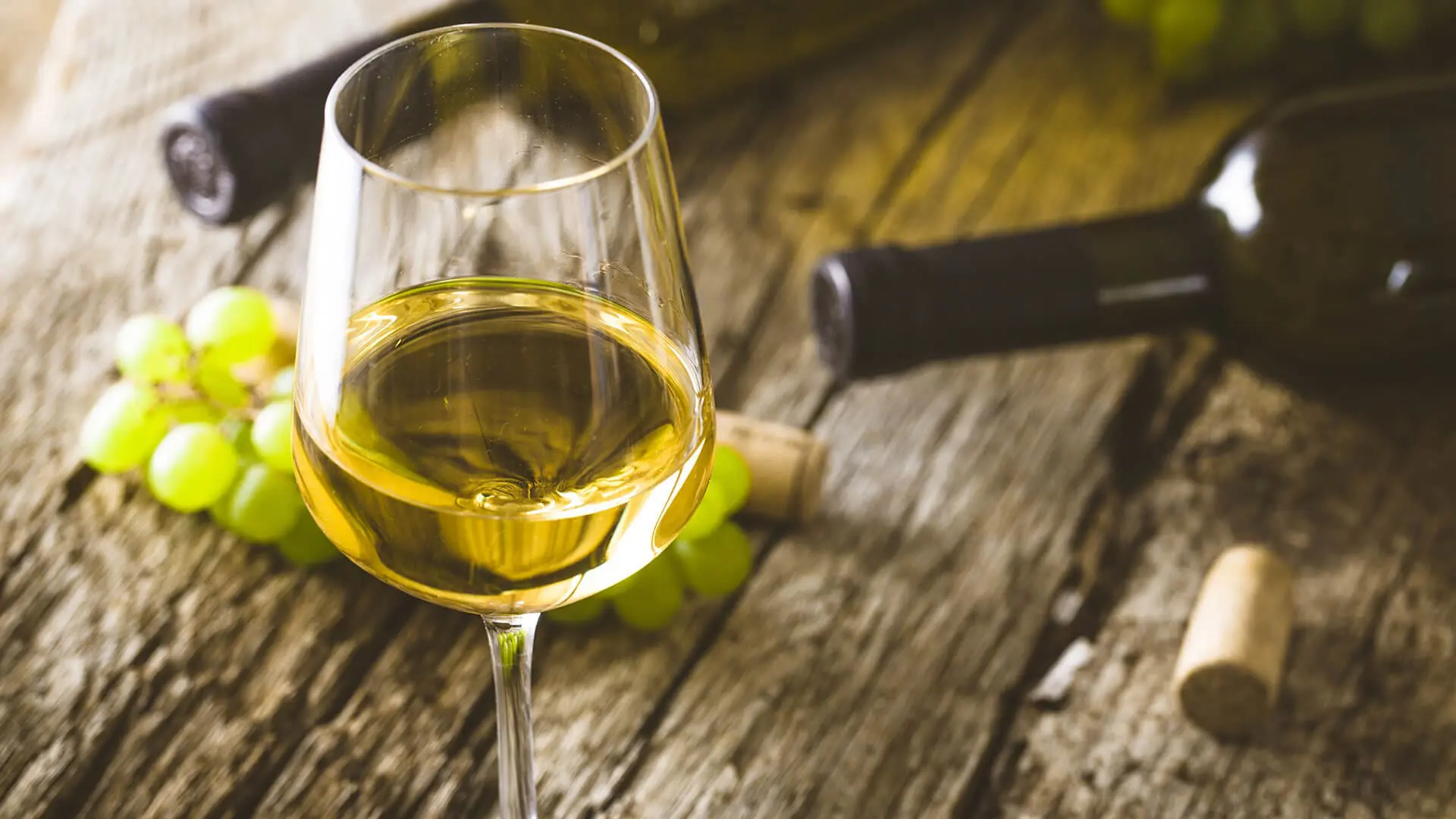 A glass of white wine on a wooden table with wine bottles, corks, and green grapes in the background evokes memories of private boat tours around Dubrovnik.