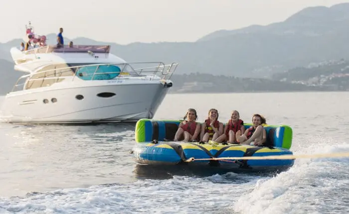 Four people wearing life jackets are being towed on an inflatable raft behind a boat in a scenic water area with mountains in the background. A motor yacht is visible nearby, likely part of one of Dubrovnik's popular private boat tours offering unforgettable day trips.