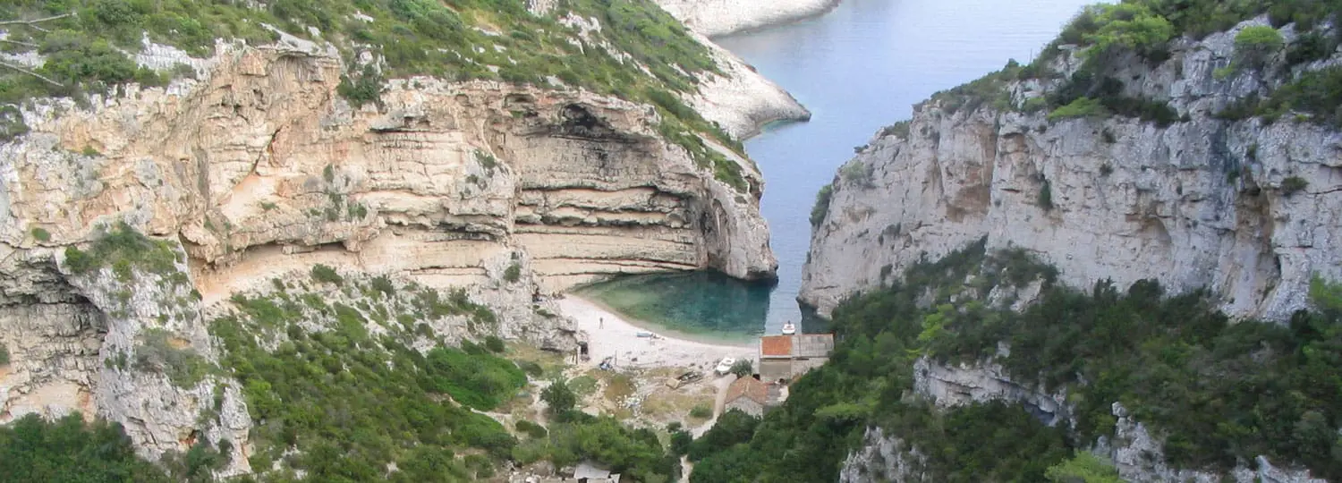 A secluded, narrow cove with clear blue water surrounded by high, rocky cliffs and lush greenery awaits you on one of our private boat tours from Dubrovnik. A small building with a red roof sits quaintly near the water's edge, perfect for those idyllic day trips.