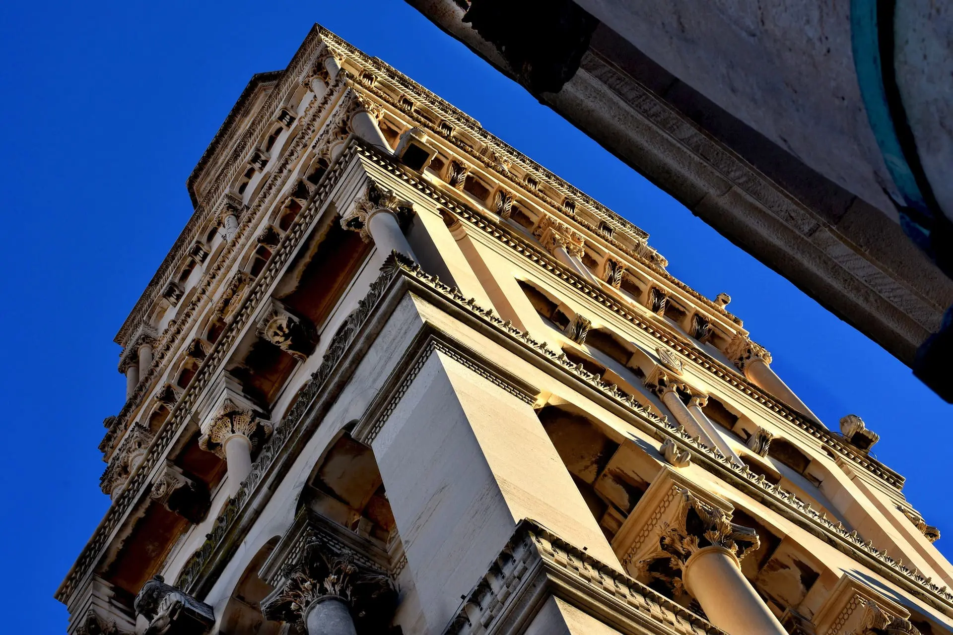 Upward view of an ornate, multi-story stone tower featuring columns and intricate carvings under a clear blue sky, reminiscent of the historic architecture seen in Dubrovnik.