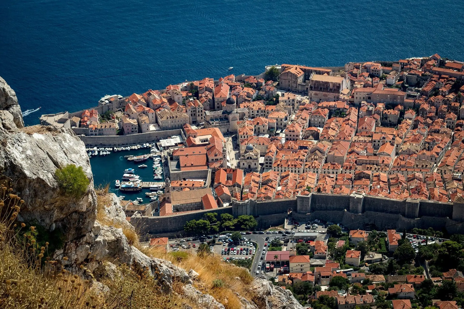 Aerial view of Dubrovnik, a coastal town with numerous orange-roofed buildings, a marina with boats, and a stone wall surrounding the area, set against a backdrop of the sea. Perfect for day trips and private boat tours.