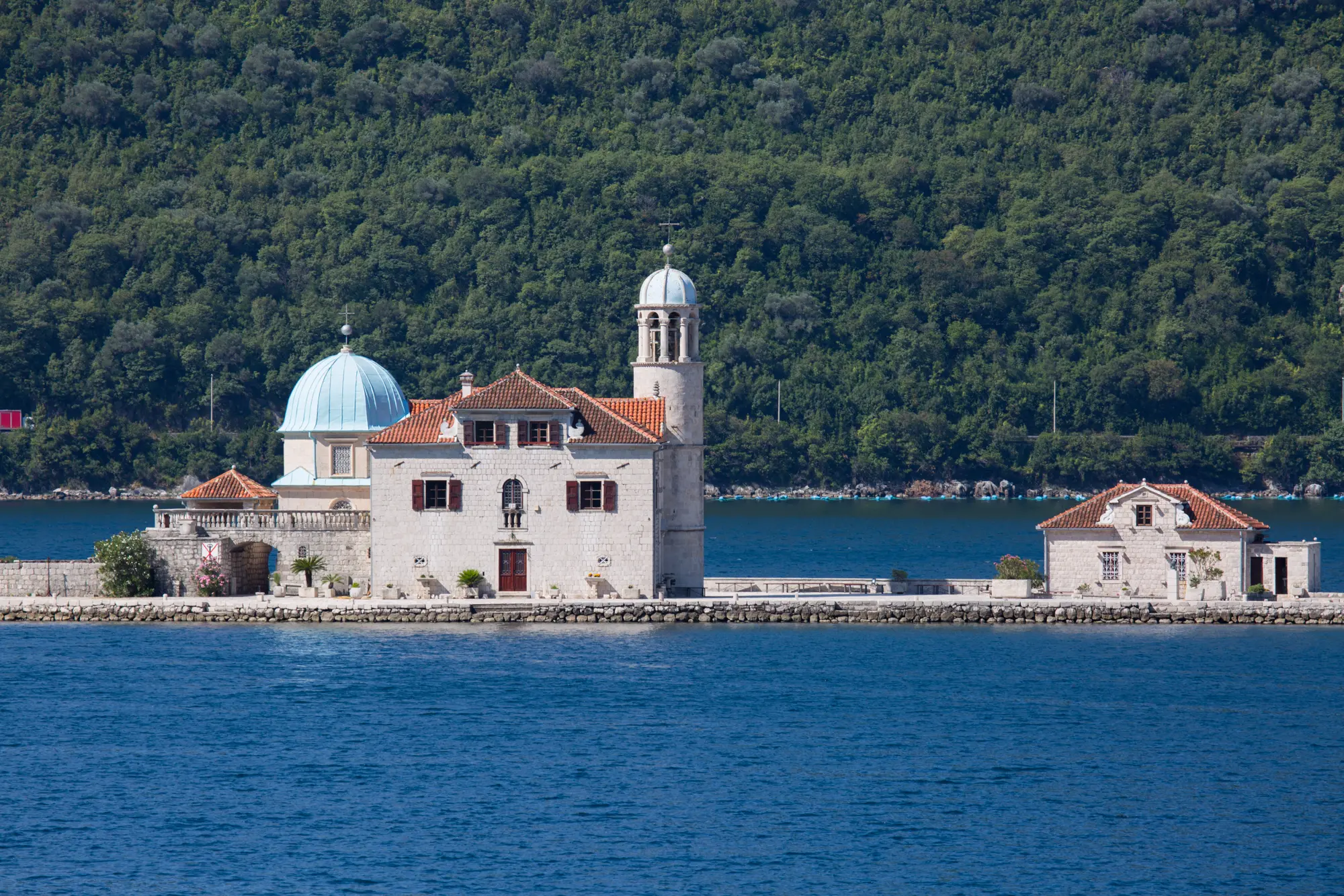 A stone building with a dome and bell tower sits on a small island surrounded by the blue waters of the Adriatic, with a forested hillside in the background, capturing the serene beauty reminiscent of Croatia's stunning landscapes.