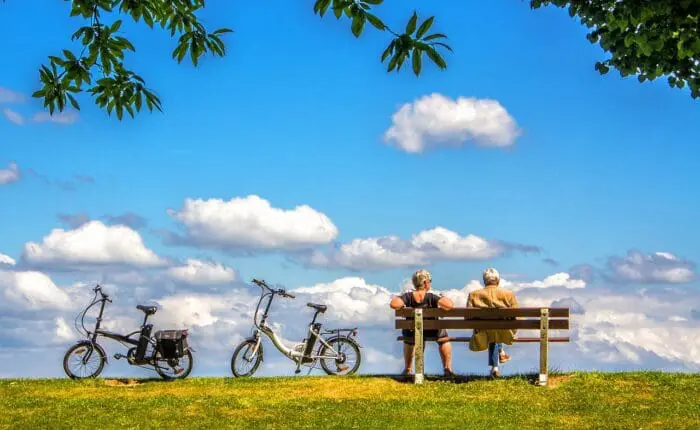 Two people sit on a bench under a blue sky with clouds in Dubrovnik, with two bicycles parked nearby.