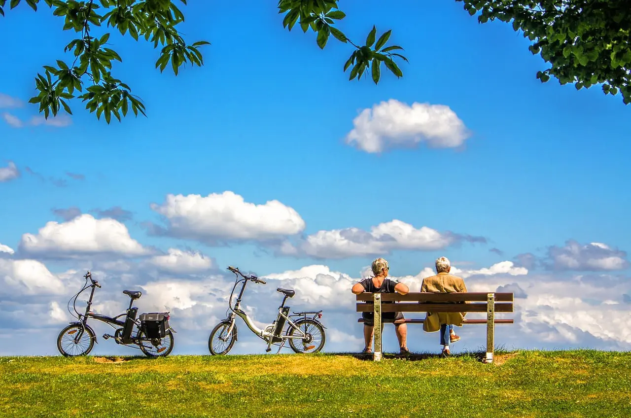 Two people sit on a bench under a blue sky with clouds in Dubrovnik, with two bicycles parked nearby.