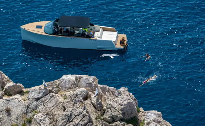 A boat is anchored near a rocky coastline in clear blue water during one of the idyllic private boat tours in Dubrovnik. Two people are swimming beside the boat while seagulls sit on the rocks.