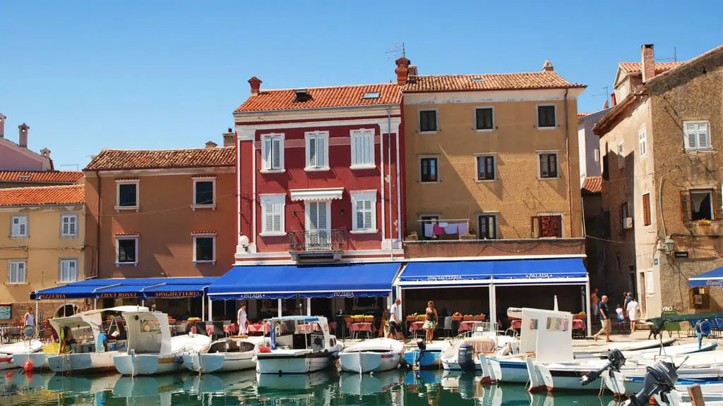 A row of colorful buildings with red-tiled roofs face a small marina with several docked boats under a clear blue sky. People walk along the waterfront and some boats have canopies.