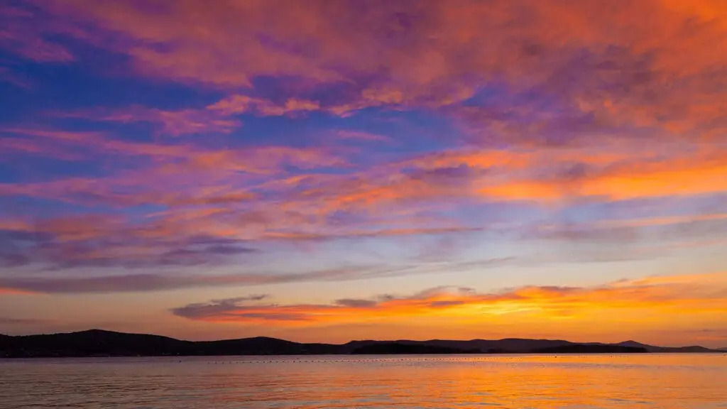 Orange, pink, and purple hues fill the sky over a calm body of water during sunset, with dark silhouettes of distant hills on the horizon.