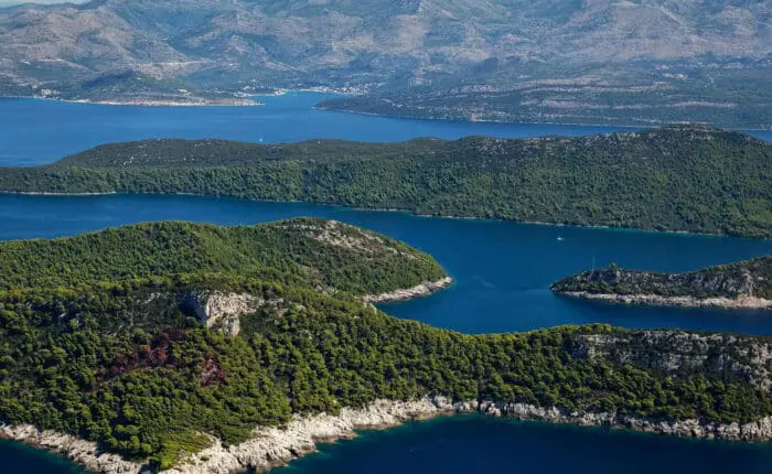 Aerial view of lush green islands surrounded by blue sea, with more islands and a mountainous landscape in the background.