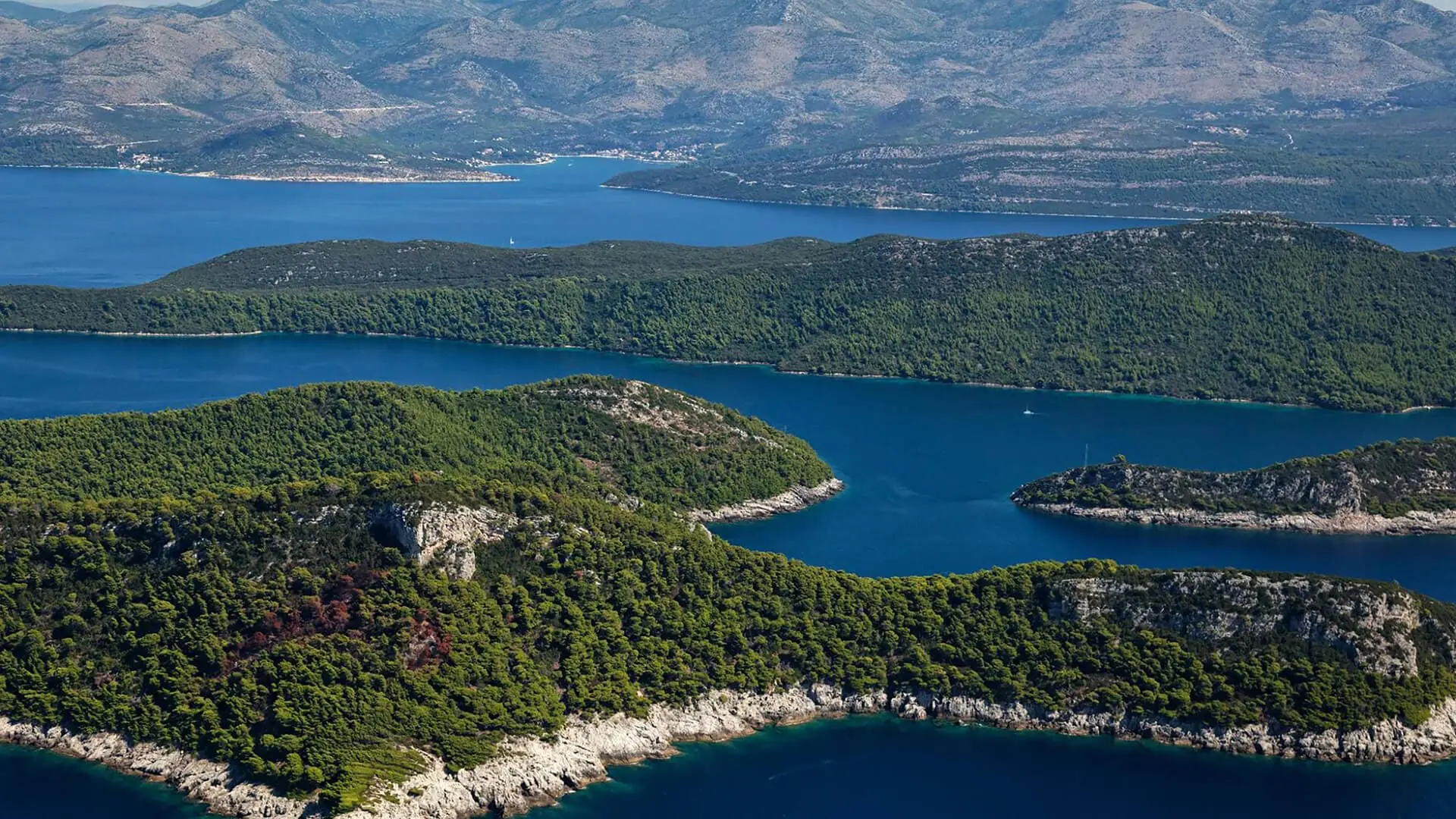 Aerial view of lush green islands surrounded by blue sea, with more islands and a mountainous landscape in the background.