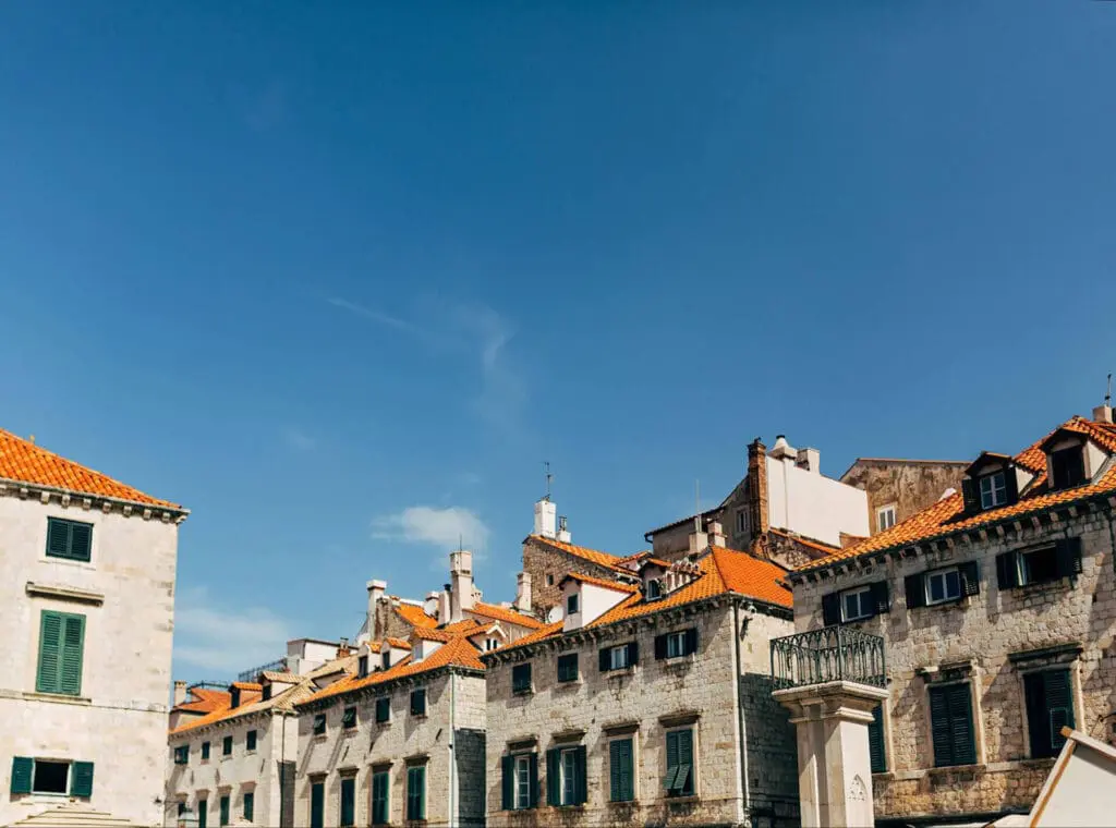 Historic stone buildings with red-tiled roofs under a clear blue sky. The architecture features small windows and a mixture of old and modern elements.