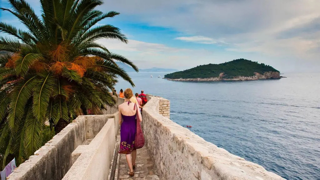 A woman in a purple dress walks along a stone pathway by the sea, with an island visible in the distance and a large palm tree nearby.