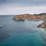 A coastal view of Dubrovnik, Croatia, showing historic walls and buildings, a few anchored boats, a small beach, and a calm sea under a cloudy sky.