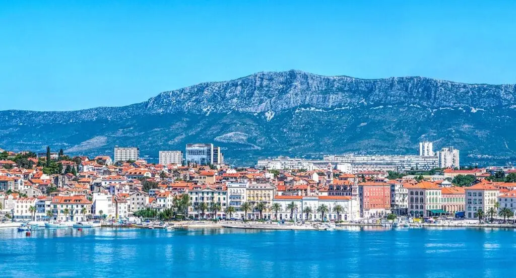 A coastal city with red-tiled roofs and modern buildings is set against a mountainous backdrop under a clear blue sky, with calm blue waters in the foreground.