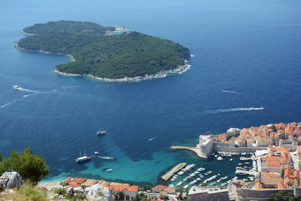 Aerial view of a coastal town with orange-roofed buildings, a marina filled with boats, and an island with lush greenery in the background.