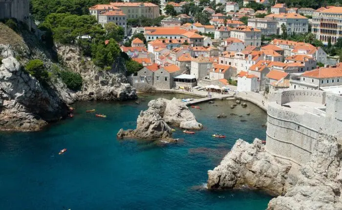 A coastal town with red-roofed buildings, surrounded by clear blue water, rocky cliffs, and kayakers in the sea.