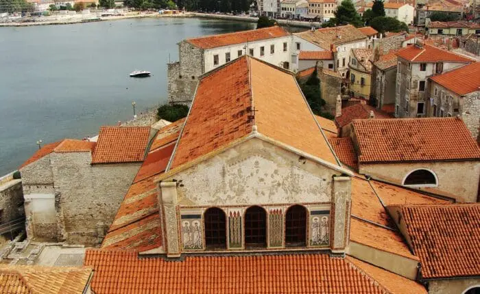 Aerial view of a coastal town in Croatia with red-tiled roofs, historic buildings, and the sparkling Adriatic Sea in the background.