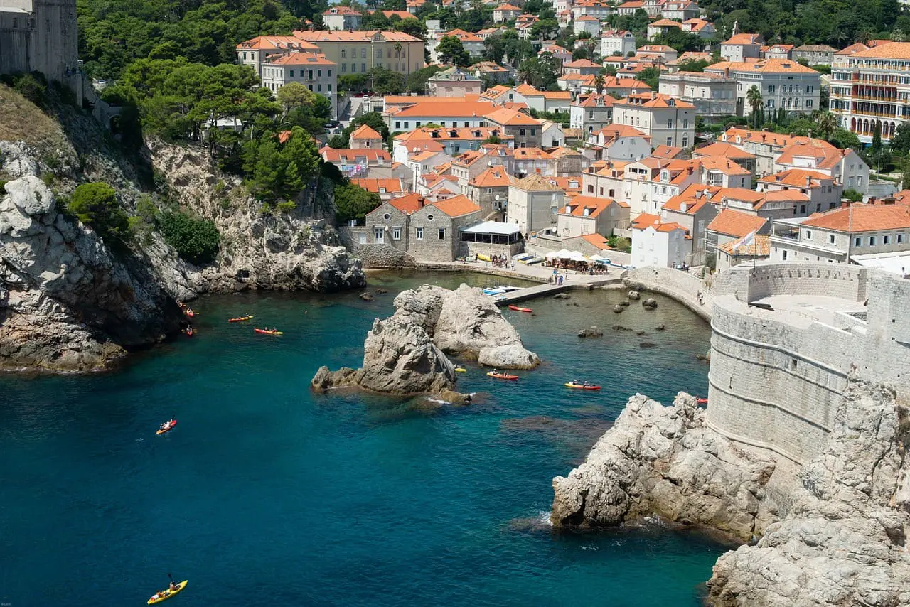 A coastal town with red-roofed buildings, surrounded by clear blue water, rocky cliffs, and kayakers in the sea.