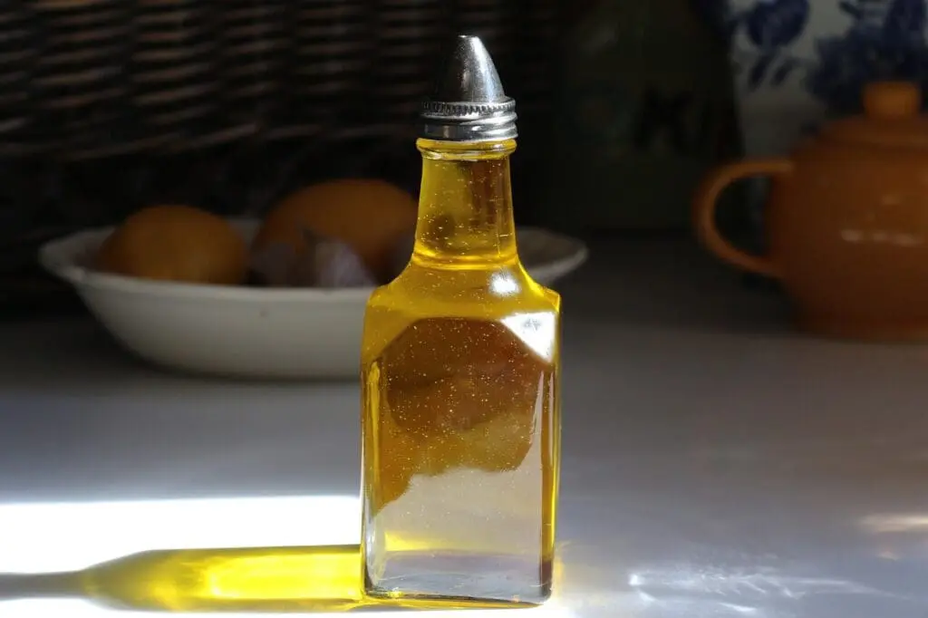 A square glass bottle filled with yellow liquid, possibly oil, with a metal pour spout, sits on a sunlit table. A bowl of lemons and a teapot are blurred in the background.