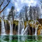 Numerous small waterfalls cascade over rocks into a clear green pool, surrounded by leafless trees on a partly cloudy day.