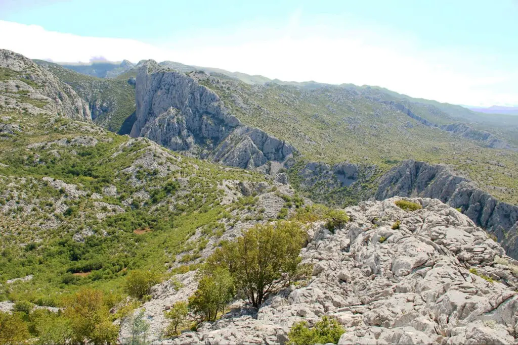 Rocky, mountainous landscape with patches of greenery and shrubs under a clear sky. The rugged terrain features steep cliffs and ridges, with distant rolling hills visible in the background.