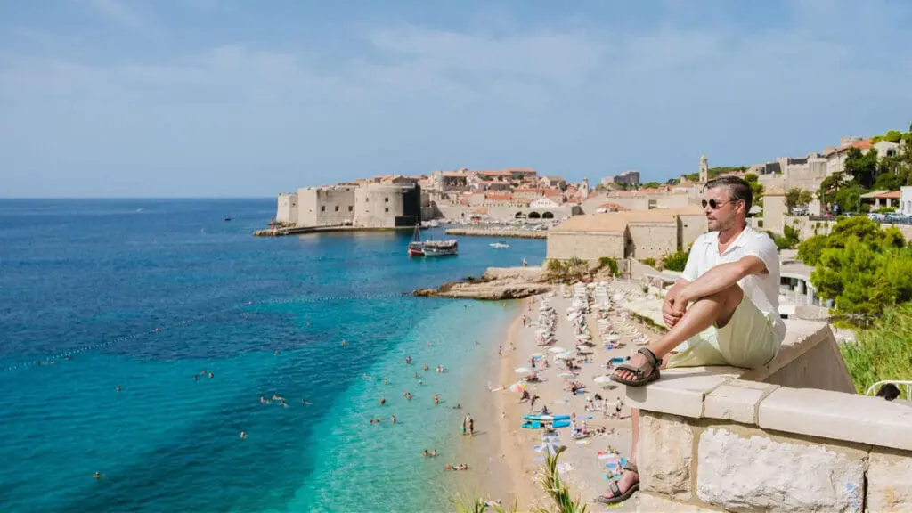 A person sitting on a stone wall overlooks a crowded beach with clear blue water and an ancient fortress in the background.