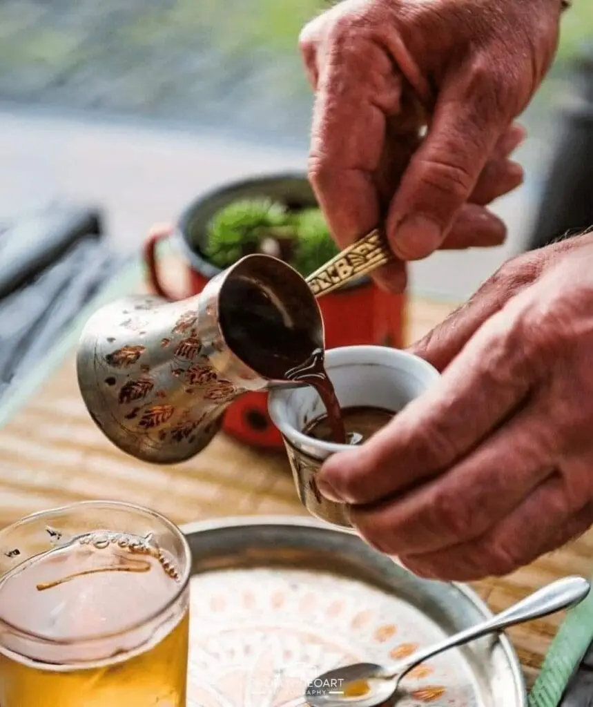 A person pours coffee from a decorated cezve into a small cup on a patterned tray, next to a glass of tea and a black pot with a plant.