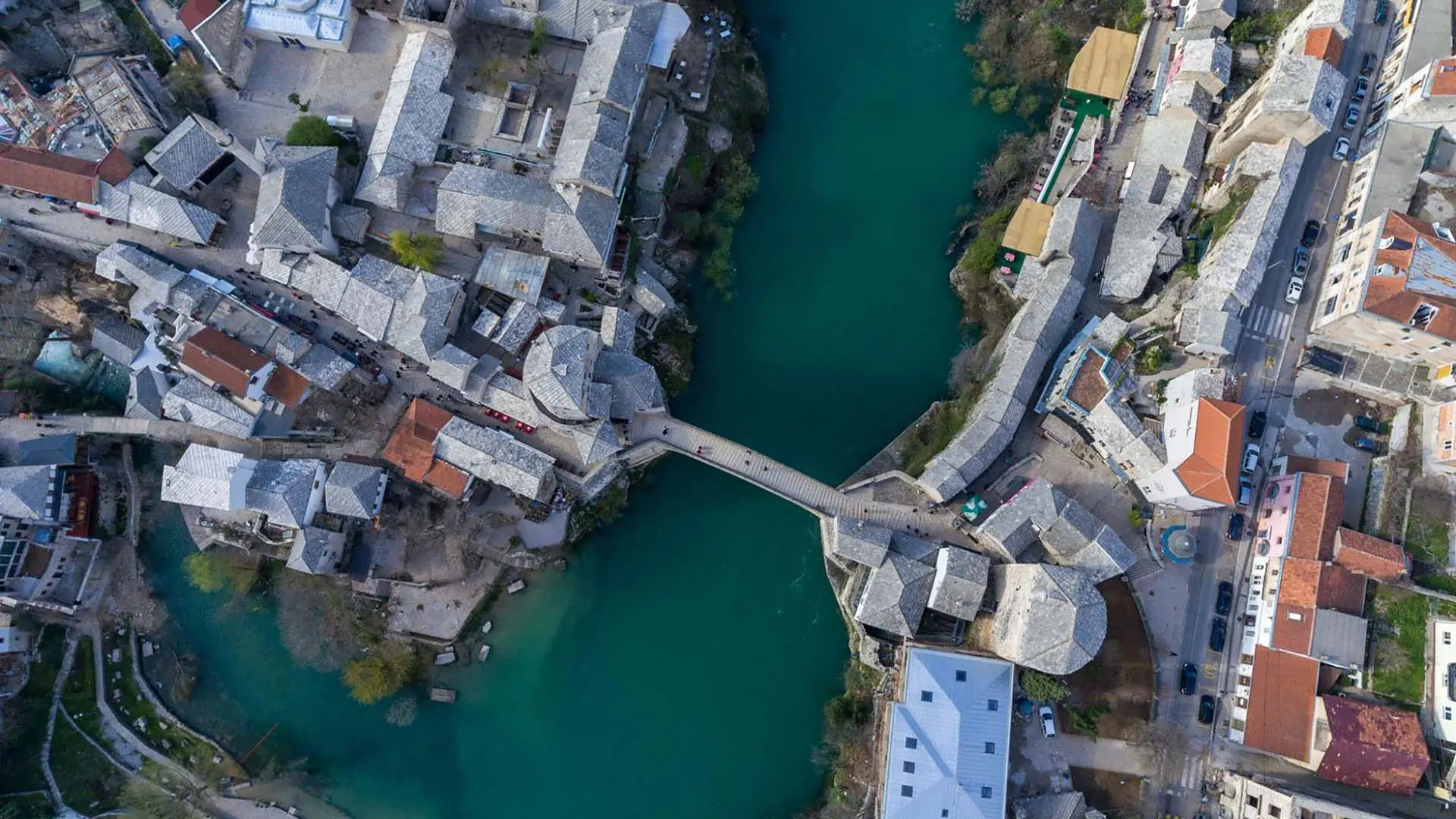 Aerial view of a bridge spanning a river, surrounded by historic stone buildings and streets. The greenish-blue water flows through the center of the town.