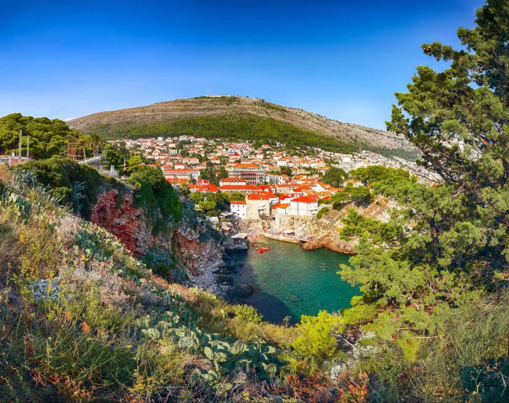 Scenic view of a coastal town with red-roofed buildings, surrounded by greenery and hills, beside clear blue water under a bright sky.