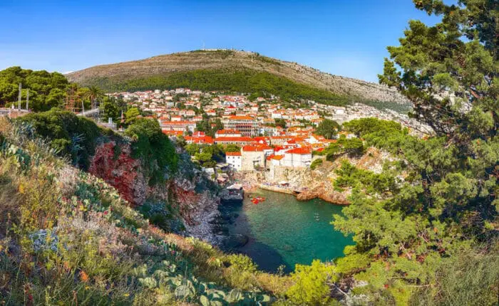 Scenic view of a coastal town with red-roofed buildings, surrounded by greenery and hills, beside clear blue water under a bright sky.