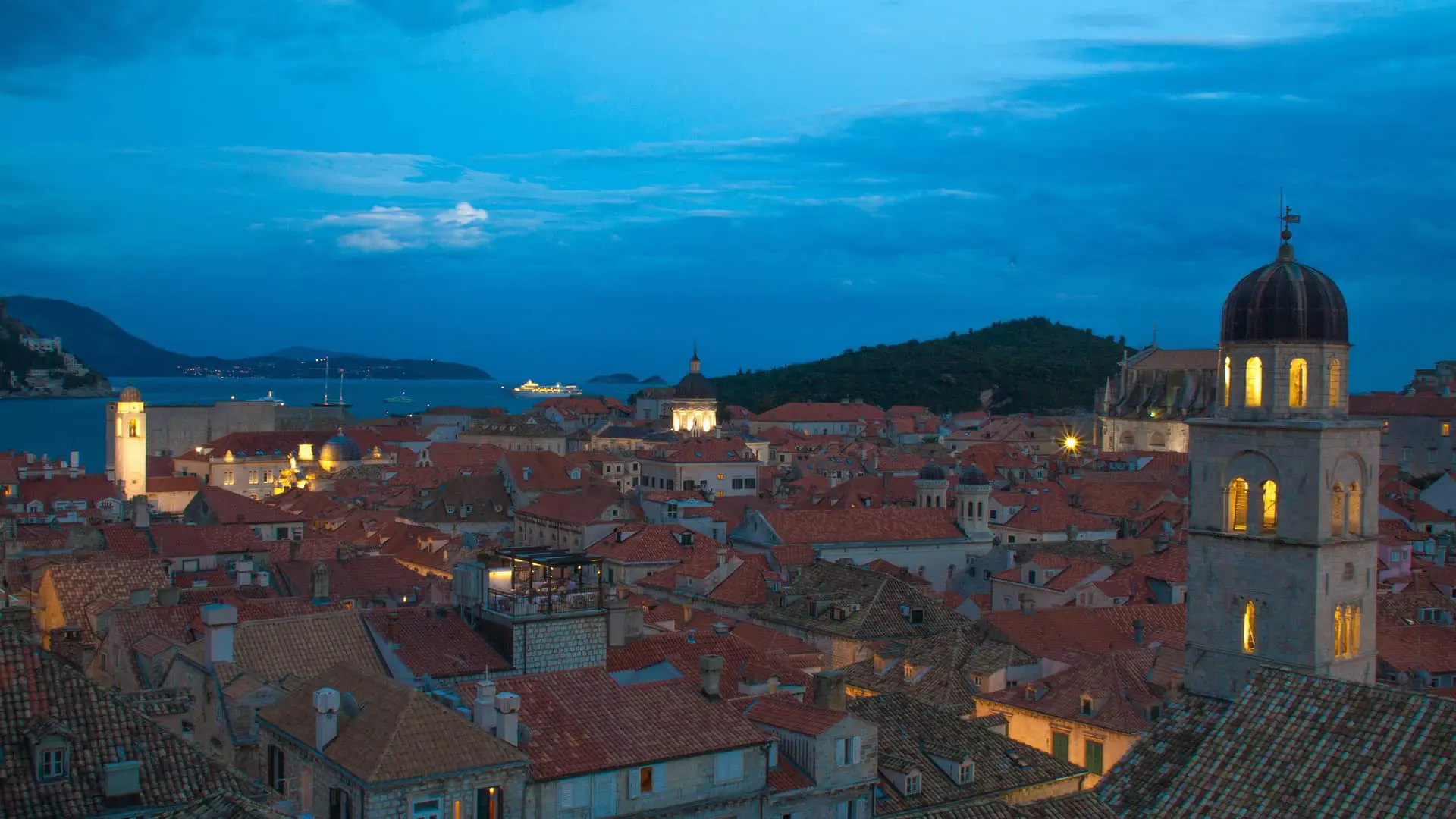 Evening view of a historic coastal city with glowing lights, red-tiled roofs, and a faintly lit church tower under a cloudy sky by the sea.