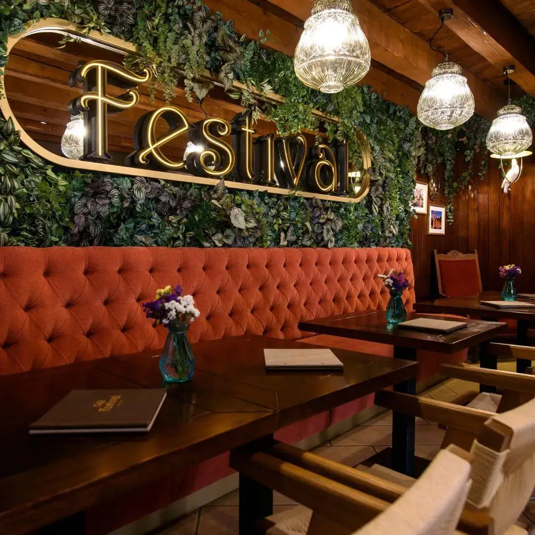 A cozy restaurant in Dubrovnik features a "Festival" sign on a greenery-covered wall, enhanced by red cushioned seating, wooden tables, and glass pendant lights. Perfect for a quiet off-season evening.