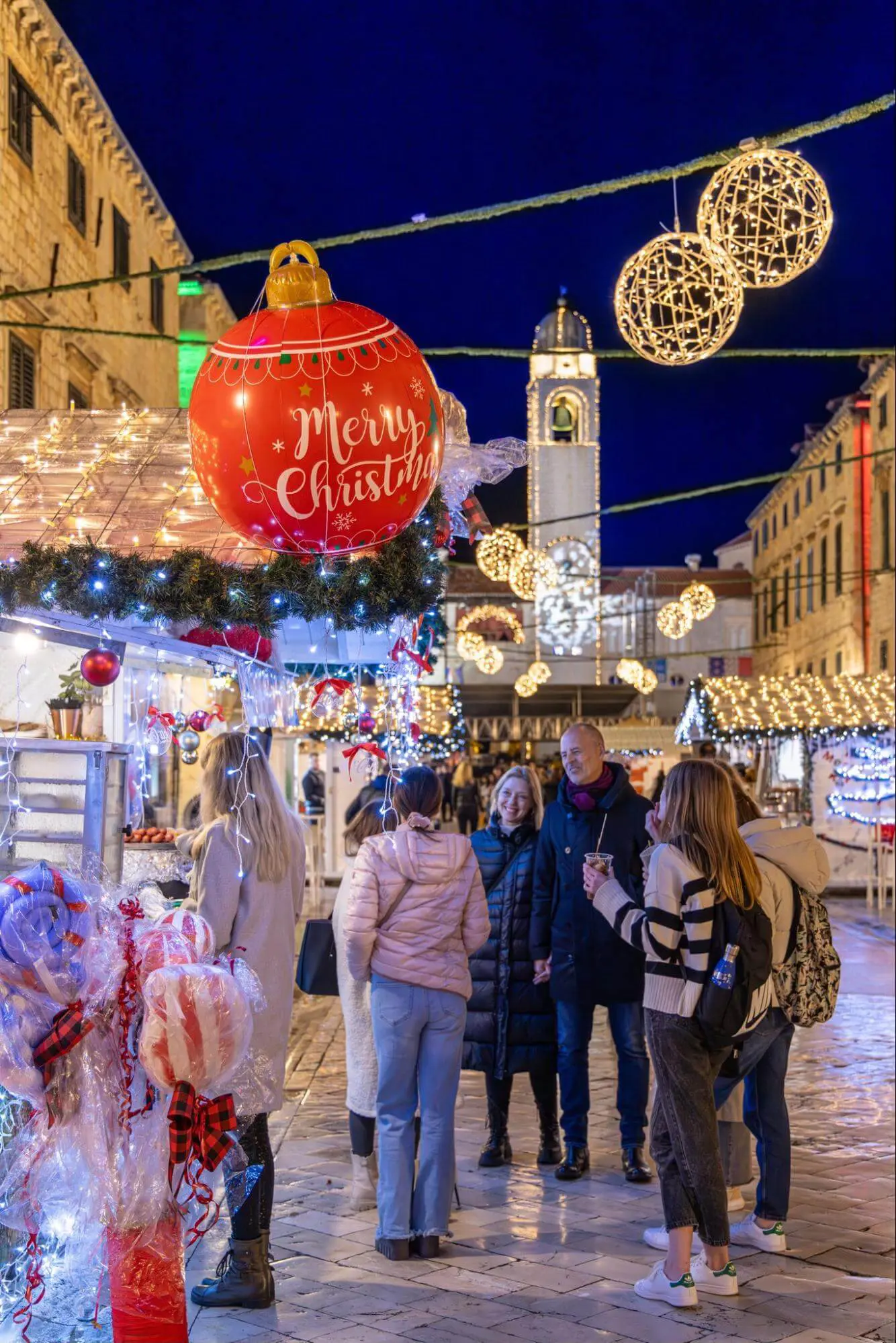 People at a Christmas market in Dubrovnik enjoy festive lights and large ornaments. A clock tower and decorated stalls sparkle under the winter's dark blue sky, capturing the charm of off-season exploration.