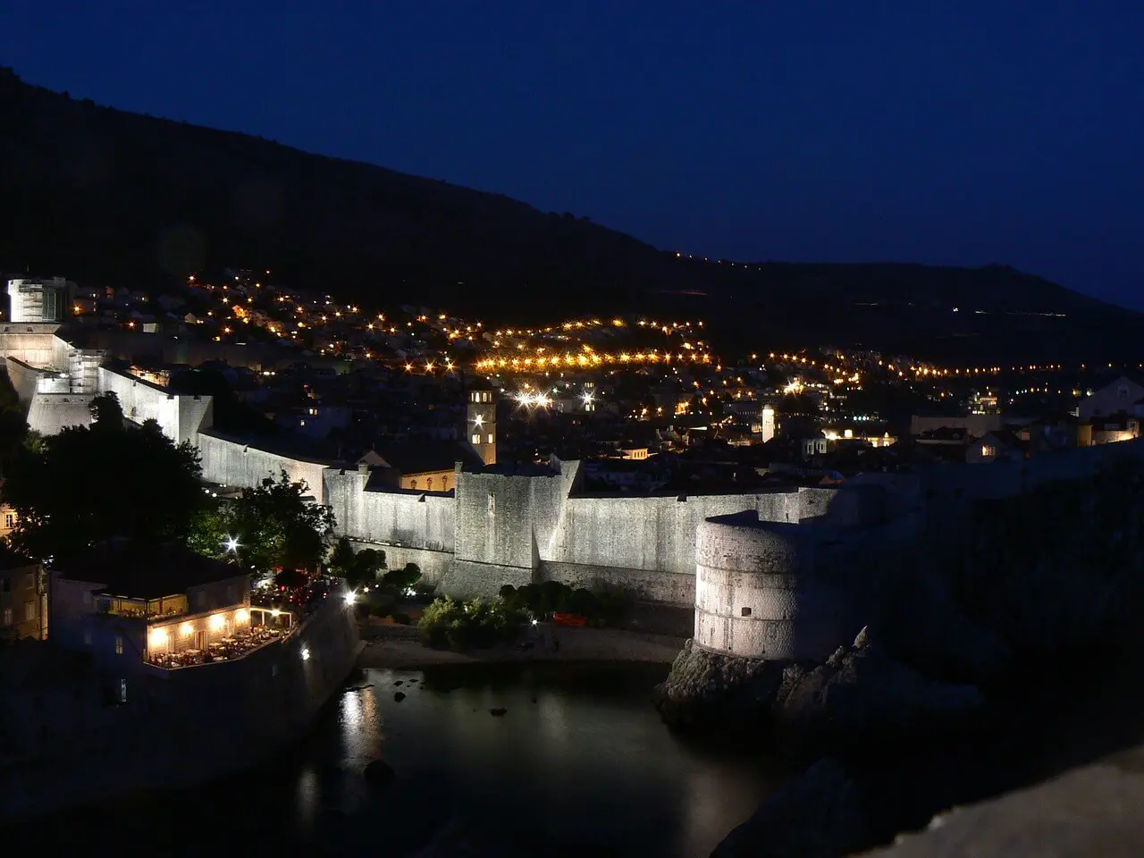 A night view of Dubrovnik's fortified stone wall surrounds the coastal town, lit by warm, glowing lights against a dark hillside backdrop, capturing the serene beauty of an off-season winter evening.