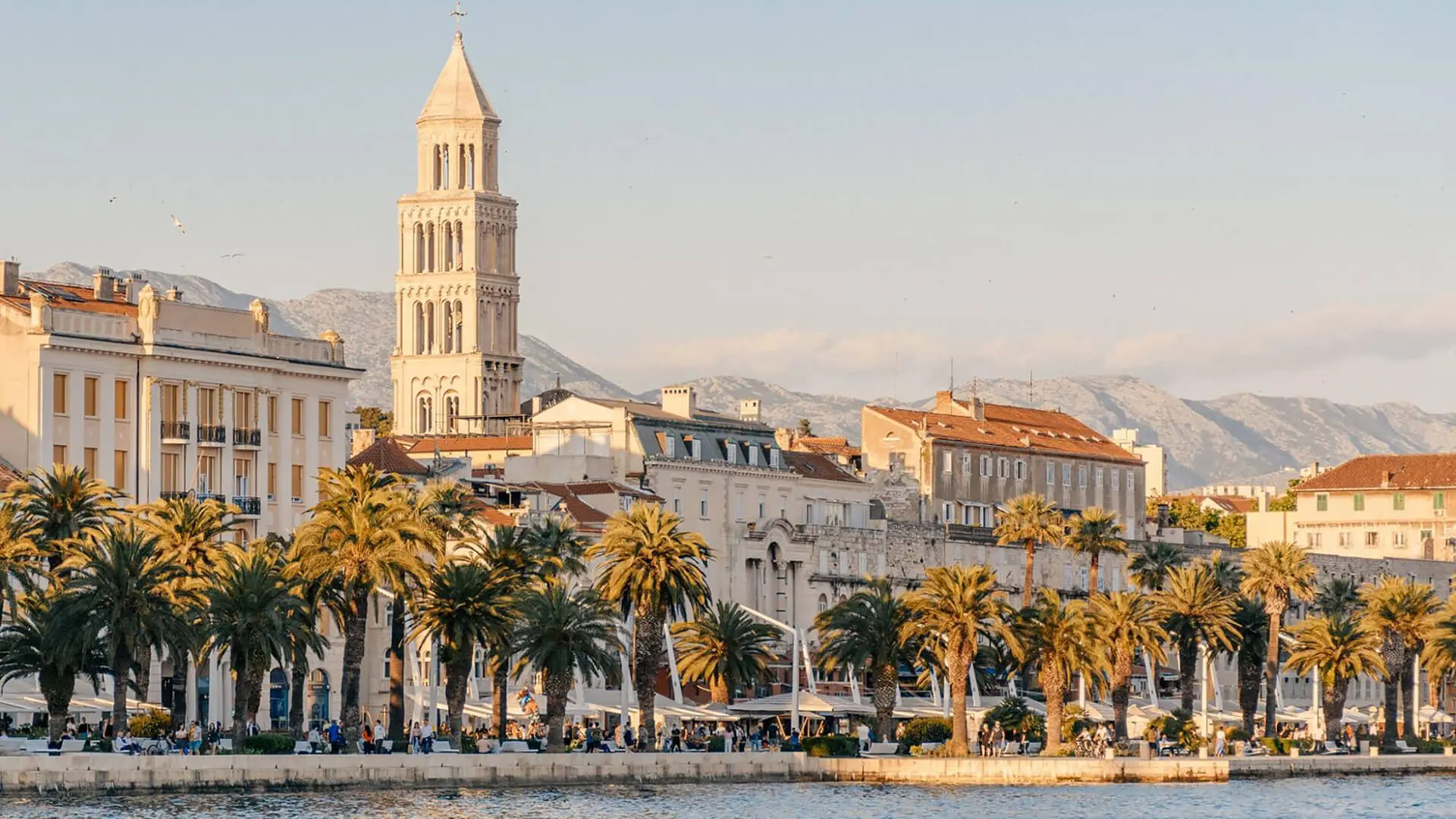 A stunning waterfront scene in Split, Croatia, with palm trees swaying by historic buildings and a tall tower. Set against the breathtaking backdrop of mountains and a clear sky, it's a perfect spring destination for travelers.