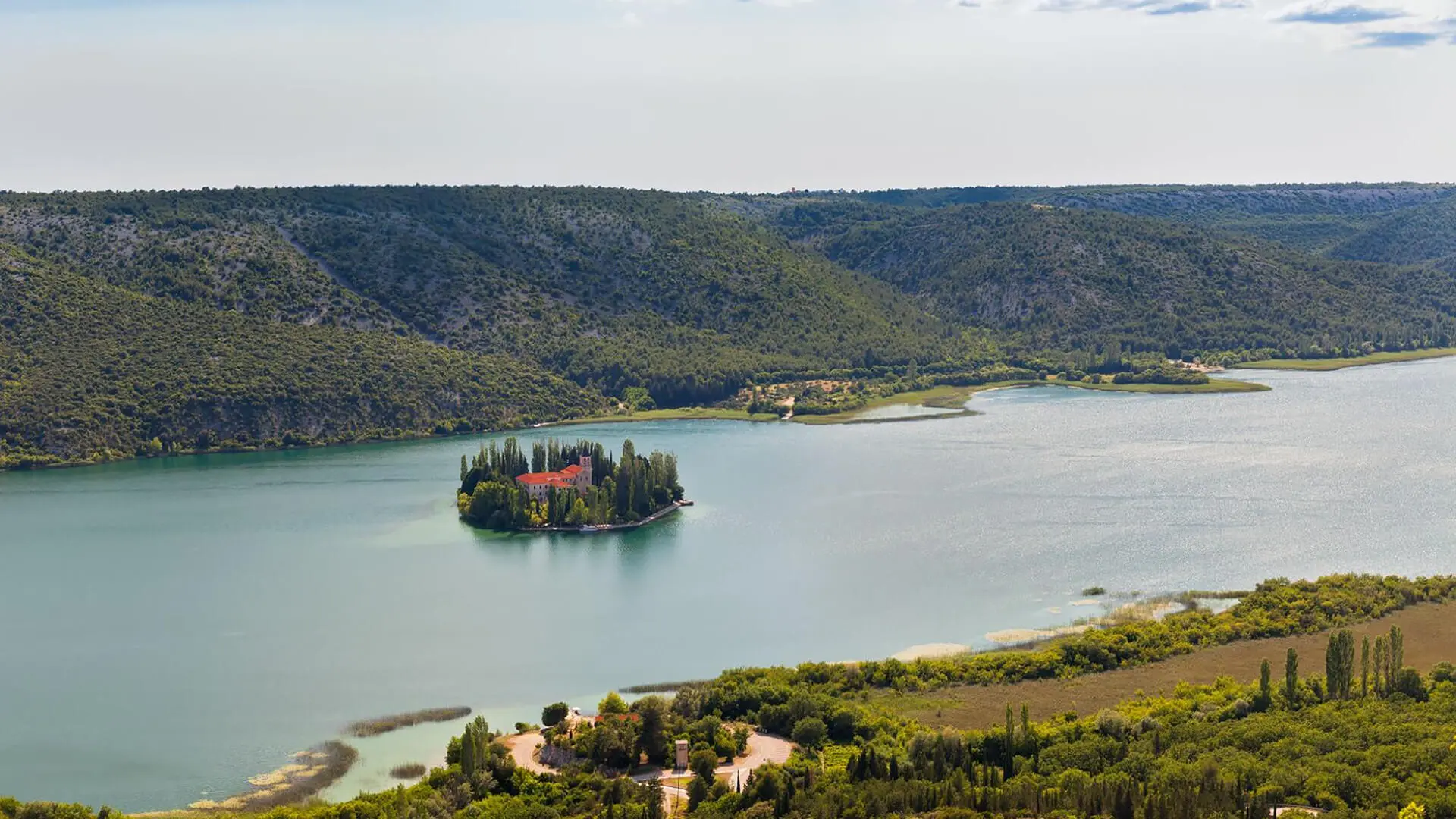 Aerial view of a small island with buildings and trees on a lake, surrounded by wooded hills—a hidden gem for travelers exploring Croatia's spring destinations.