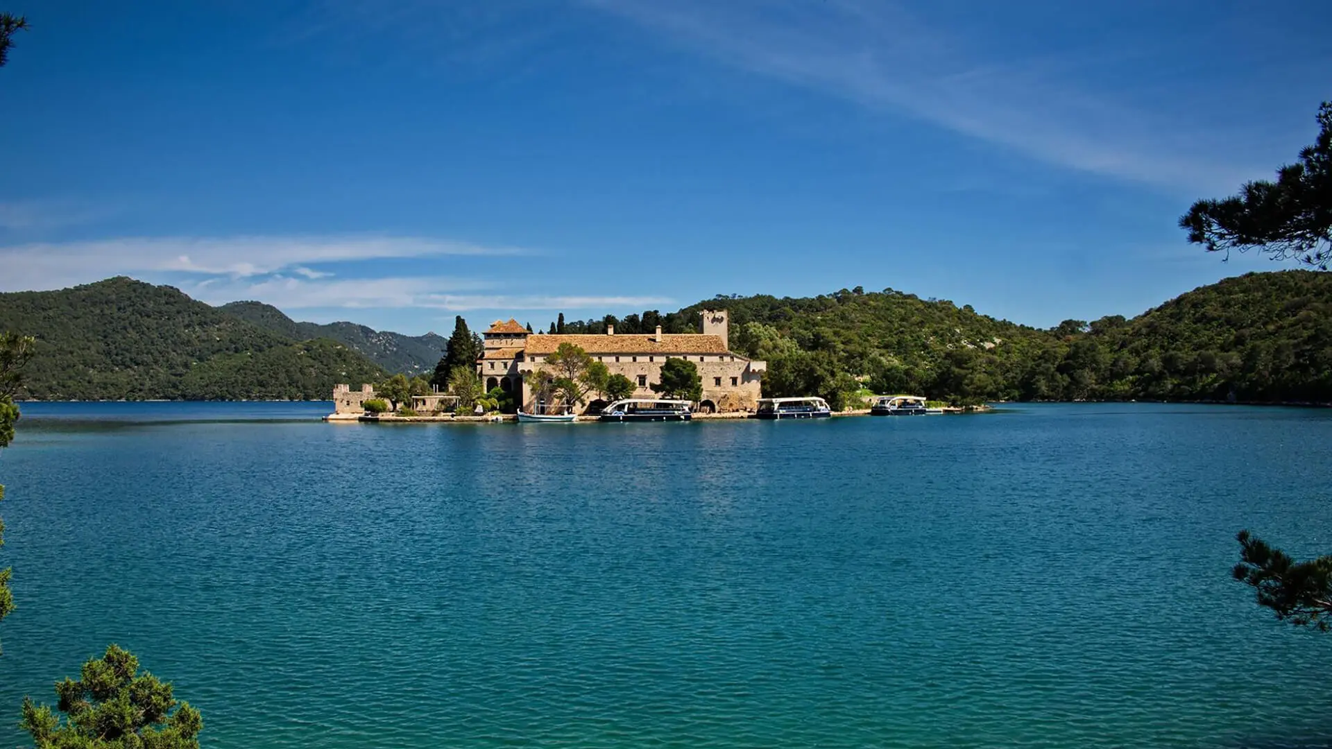 A picturesque island in Croatia, popular among spring destinations for travelers, features a historic building amidst lush greenery. Surrounded by blue water under a clear sky, with hills gracefully framing the backdrop.