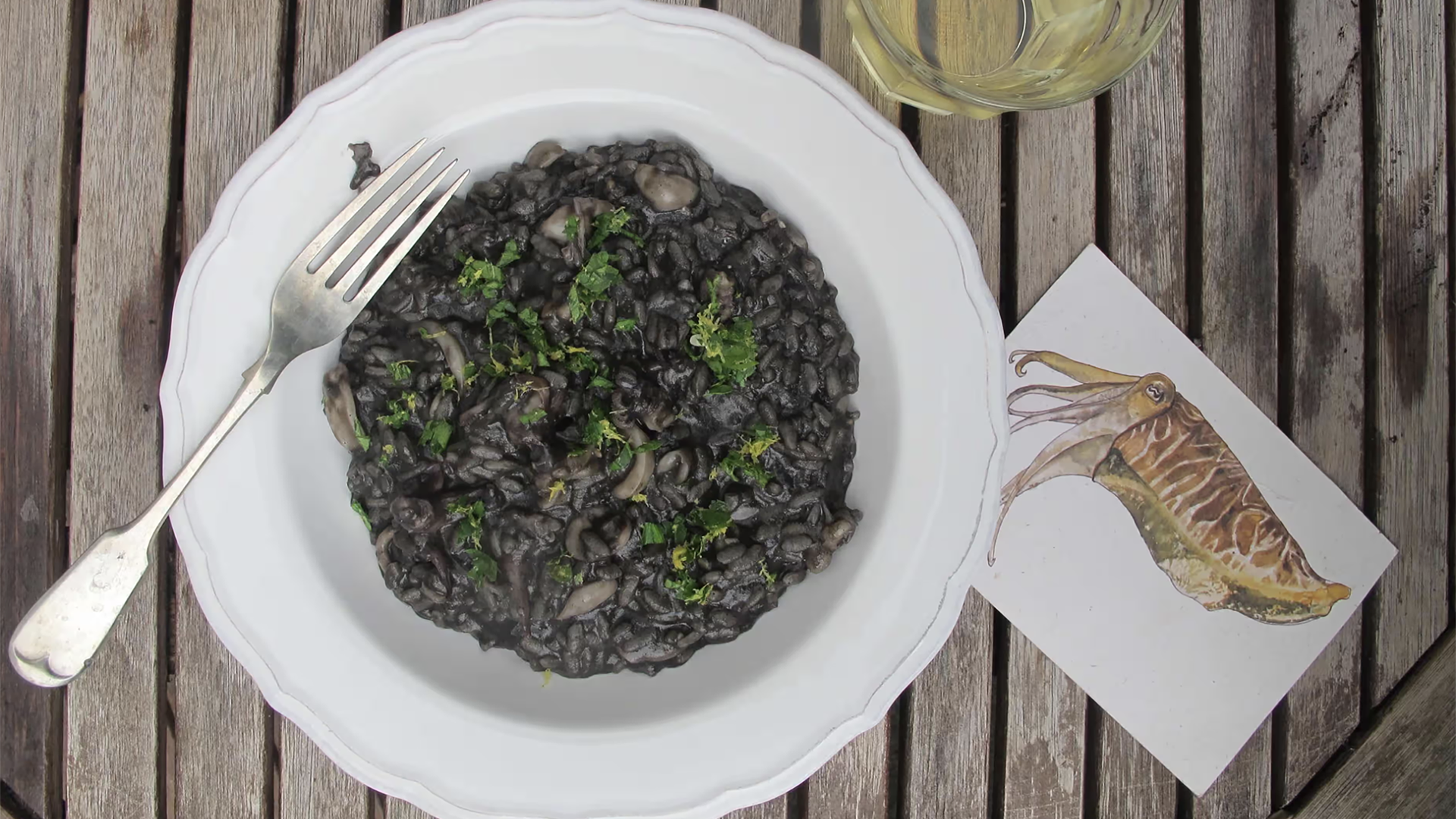 A bowl of black risotto with parsley garnish and a fork sits on a wooden table, evoking the flavors of Dalmatian dishes. Nearby, a glass of white wine complements the meal, alongside a postcard featuring a squid illustration—perfect for those traveling through culinary delights.