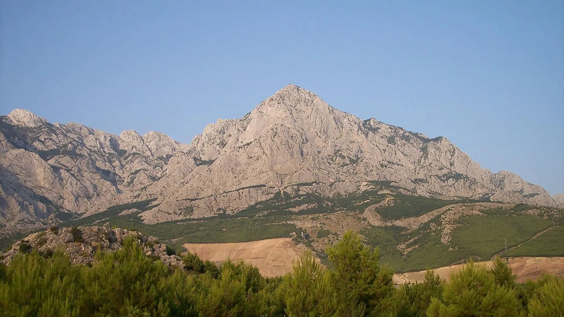 A mountain peak with rocky slopes under a clear blue sky, surrounded by green hills and trees in the foreground, invites exploration. Nearby, Croatia's natural springs offer rejuvenating wellness getaways amidst the serene beauty of nature.