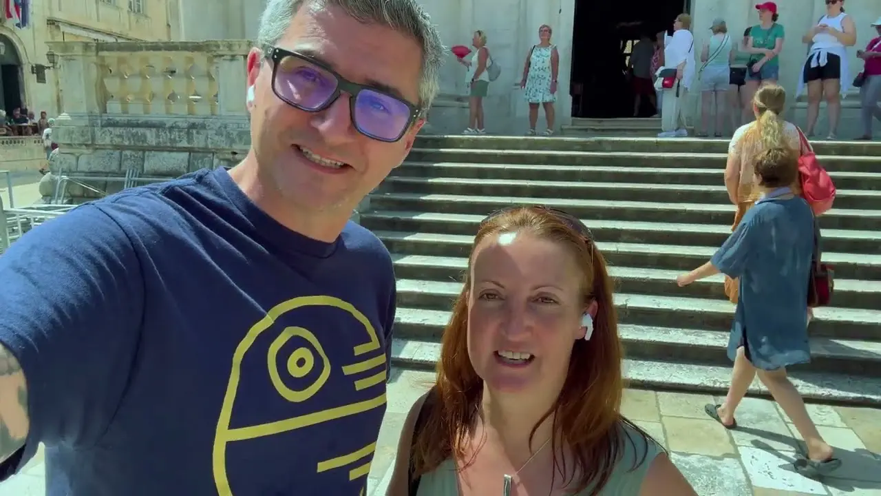 A man and a woman stand in front of stone steps outdoors, with several people in the background and a historic building behind them.