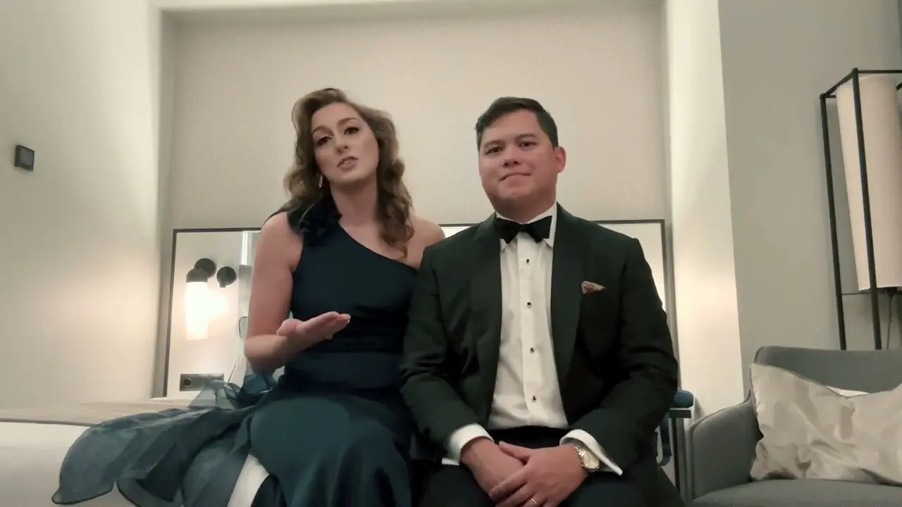 A woman in a dark one-shoulder gown and a man in a tuxedo sit side by side on a bed in a modern, well-lit room.