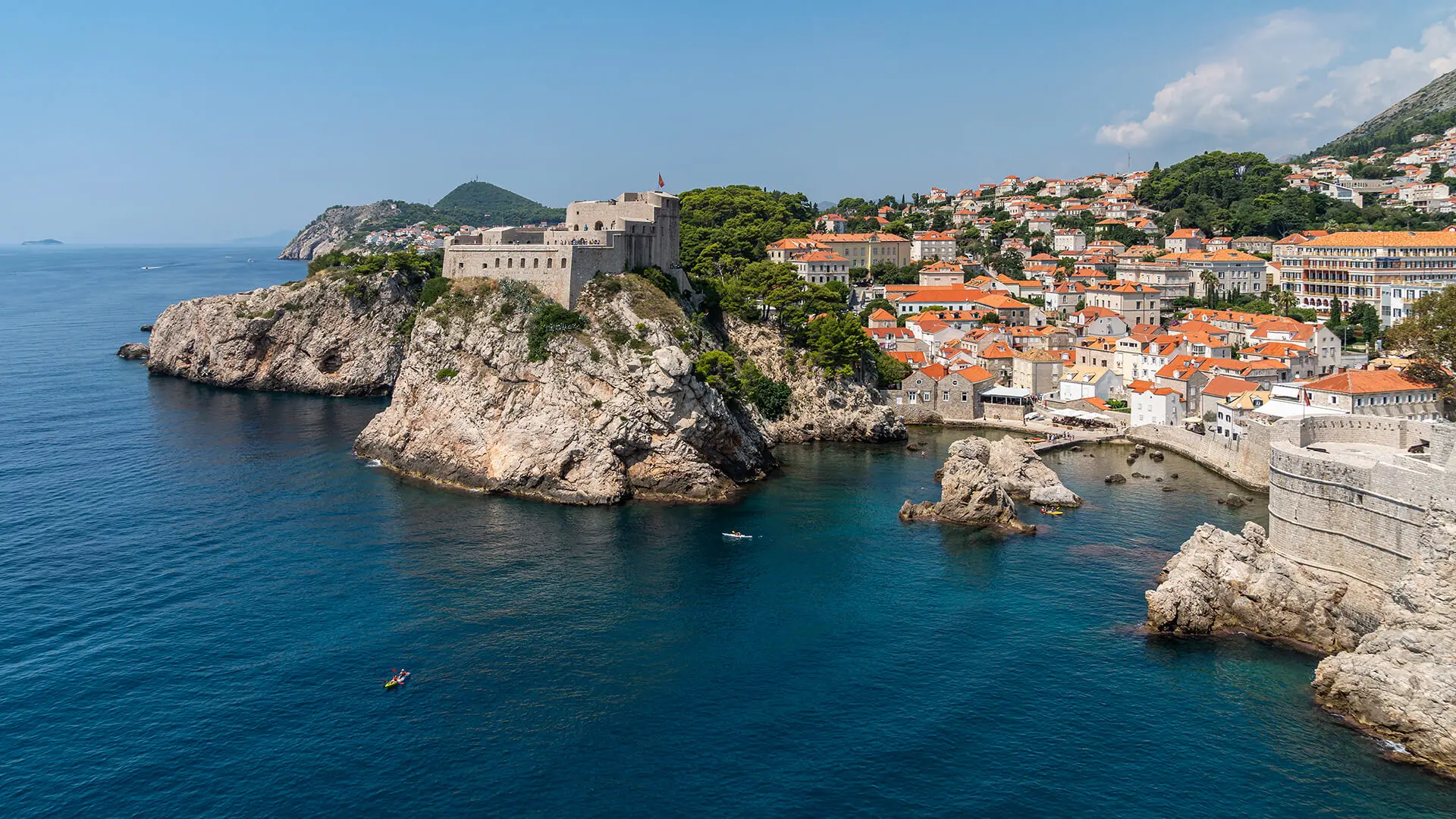 A coastal city with stone buildings and orange roofs, Dubrovnik features a large fortress on a rocky cliff and a calm blue sea—perfect for exploring things to do in July on a sunny day.