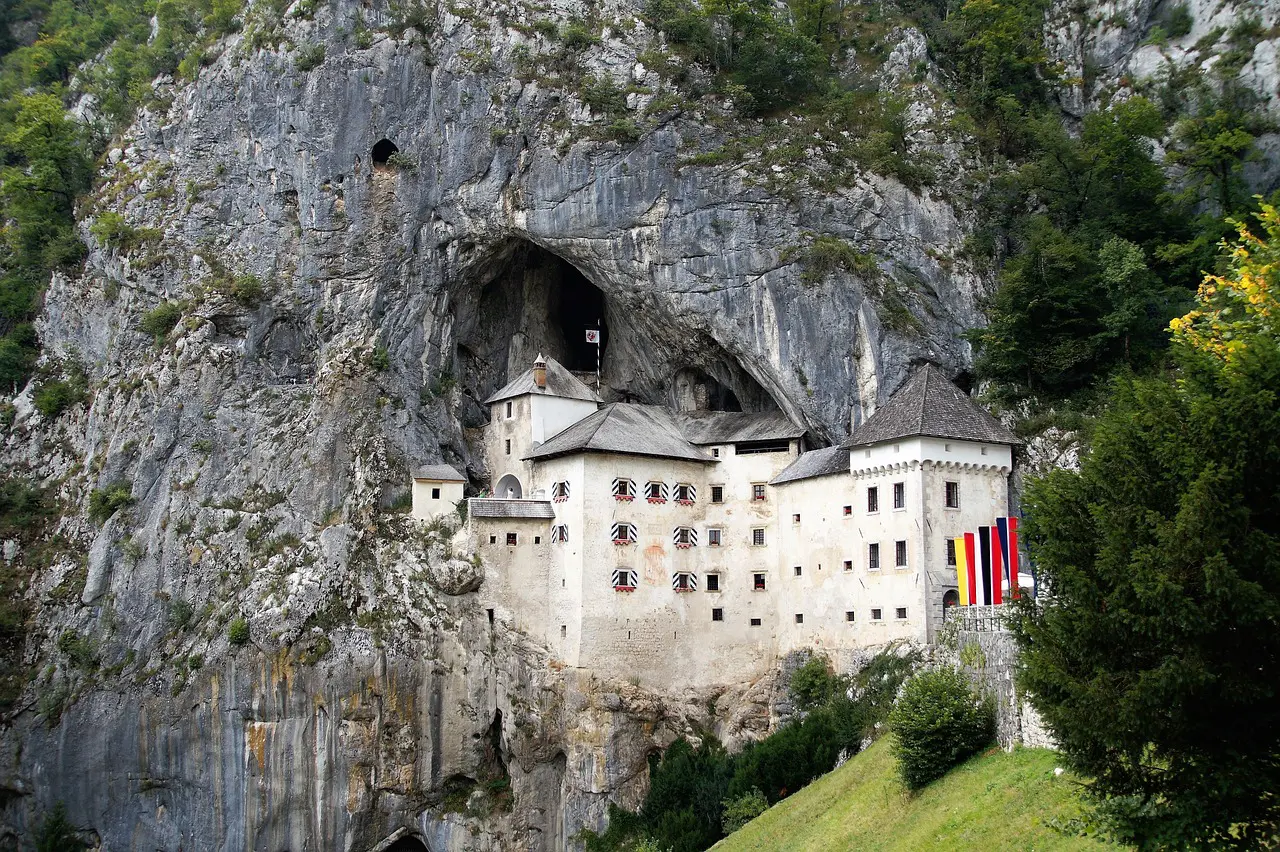 A large stone castle built into the side of a rocky cliff, with multiple windows and three vertical red, yellow, and black flags hanging outside. Trees surround the structure—a highlight on many Slovenia tours.